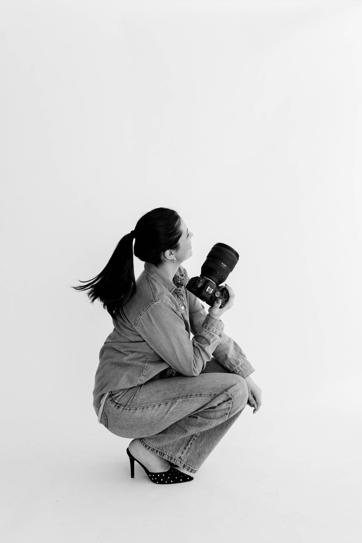 Stylish photographer in black outfit and white boots poses with camera while sitting on director's chair against white wall.