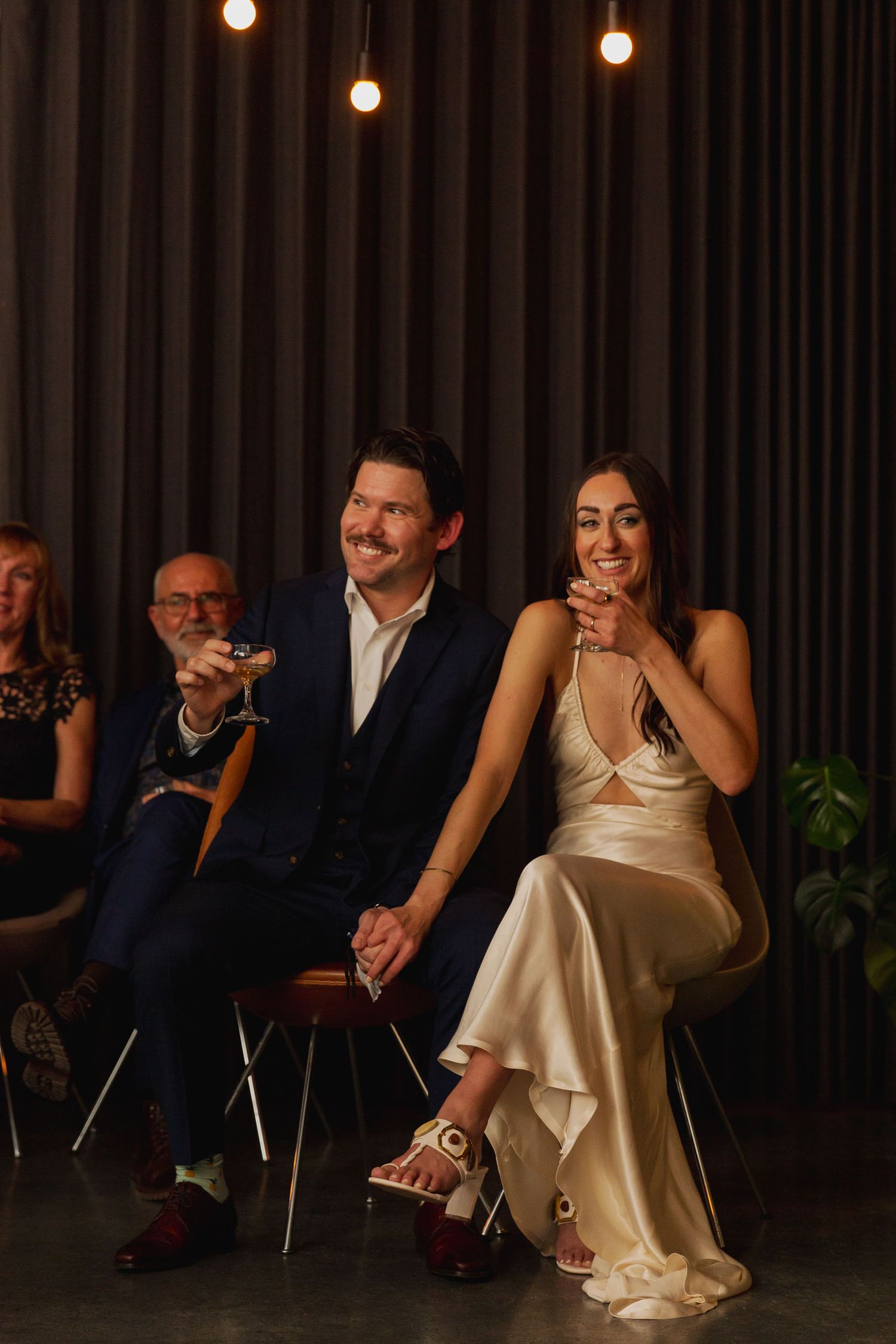 Audience members in formal attire enjoying an indoor event with warm lighting and dark curtains in the background.