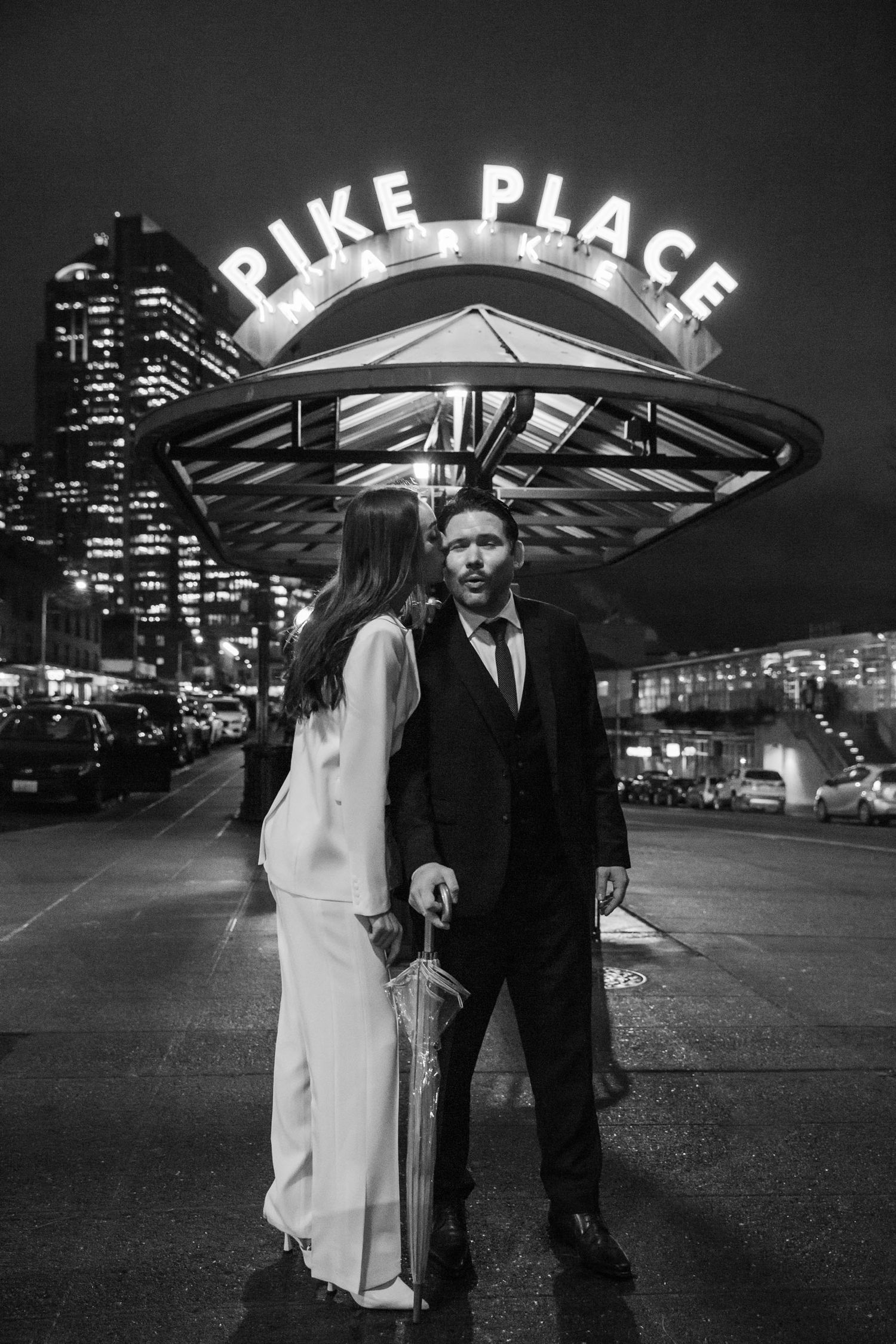 A couple shares a romantic moment under the iconic Pike Place Market sign in Seattle at night.