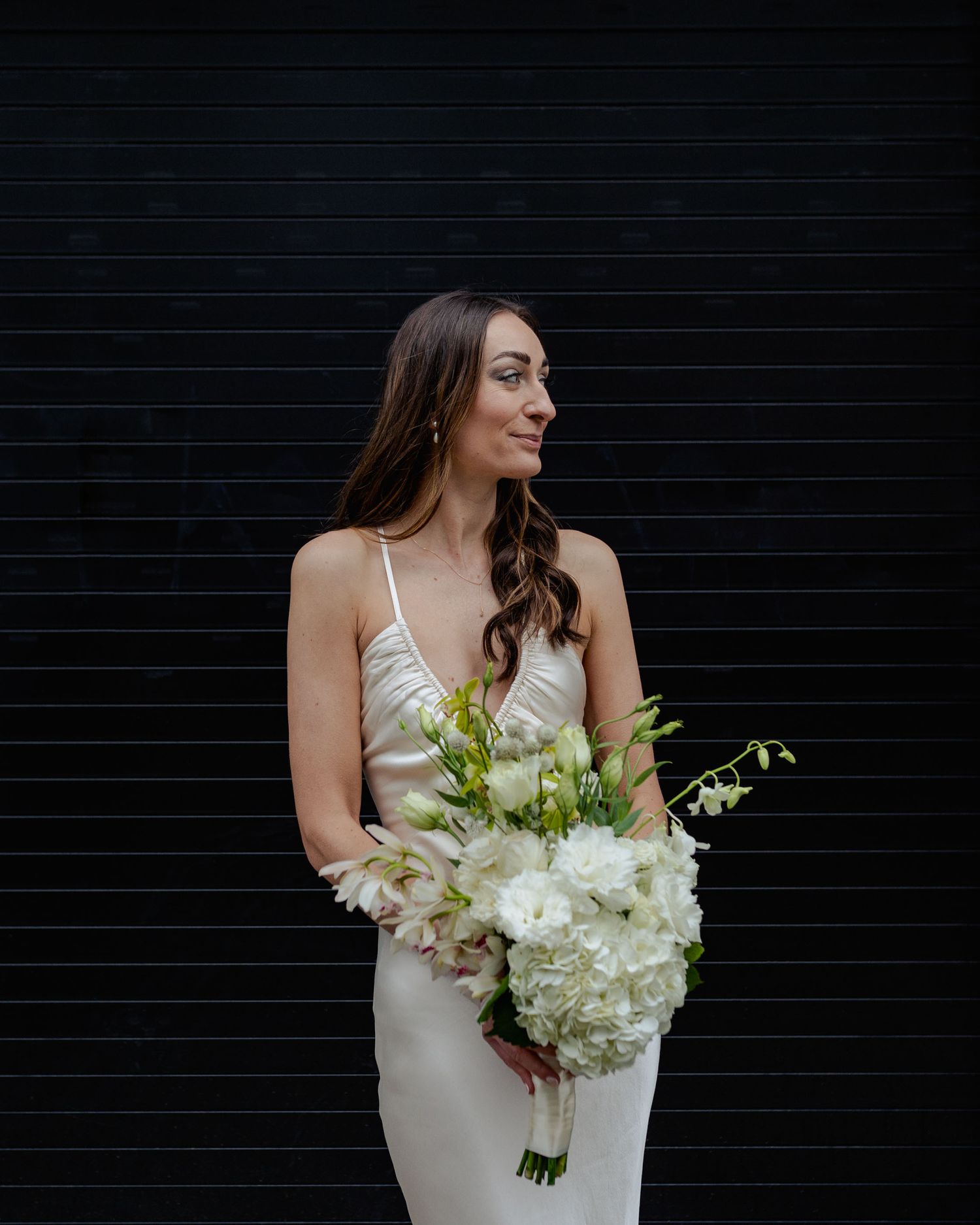 A bride in a sleek white dress holds an elegant bouquet of white hydrangeas and greenery against a black wall.