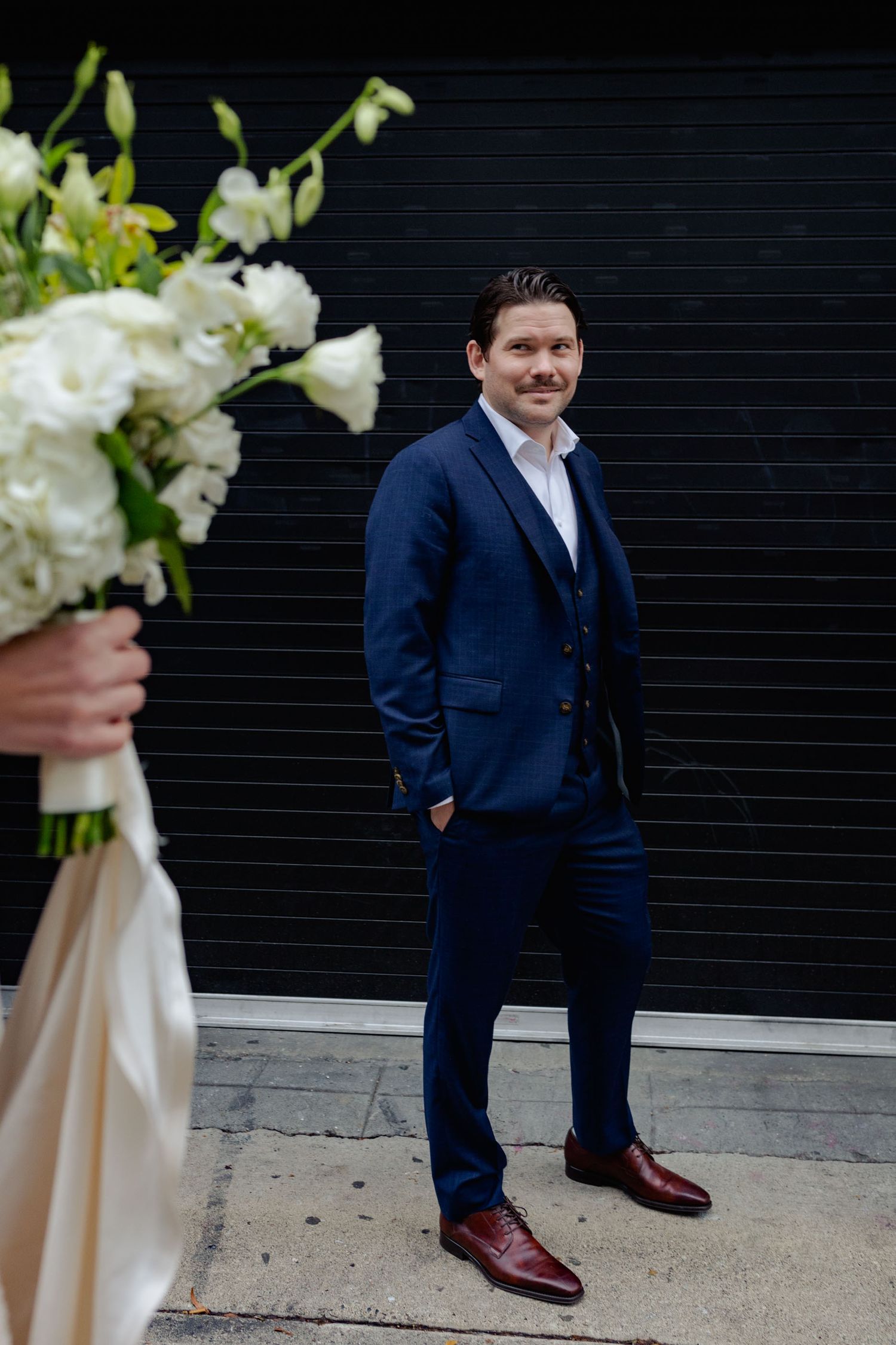 A person in a navy blue suit stands on a sidewalk next to white wedding bouquet flowers against a dark wall.