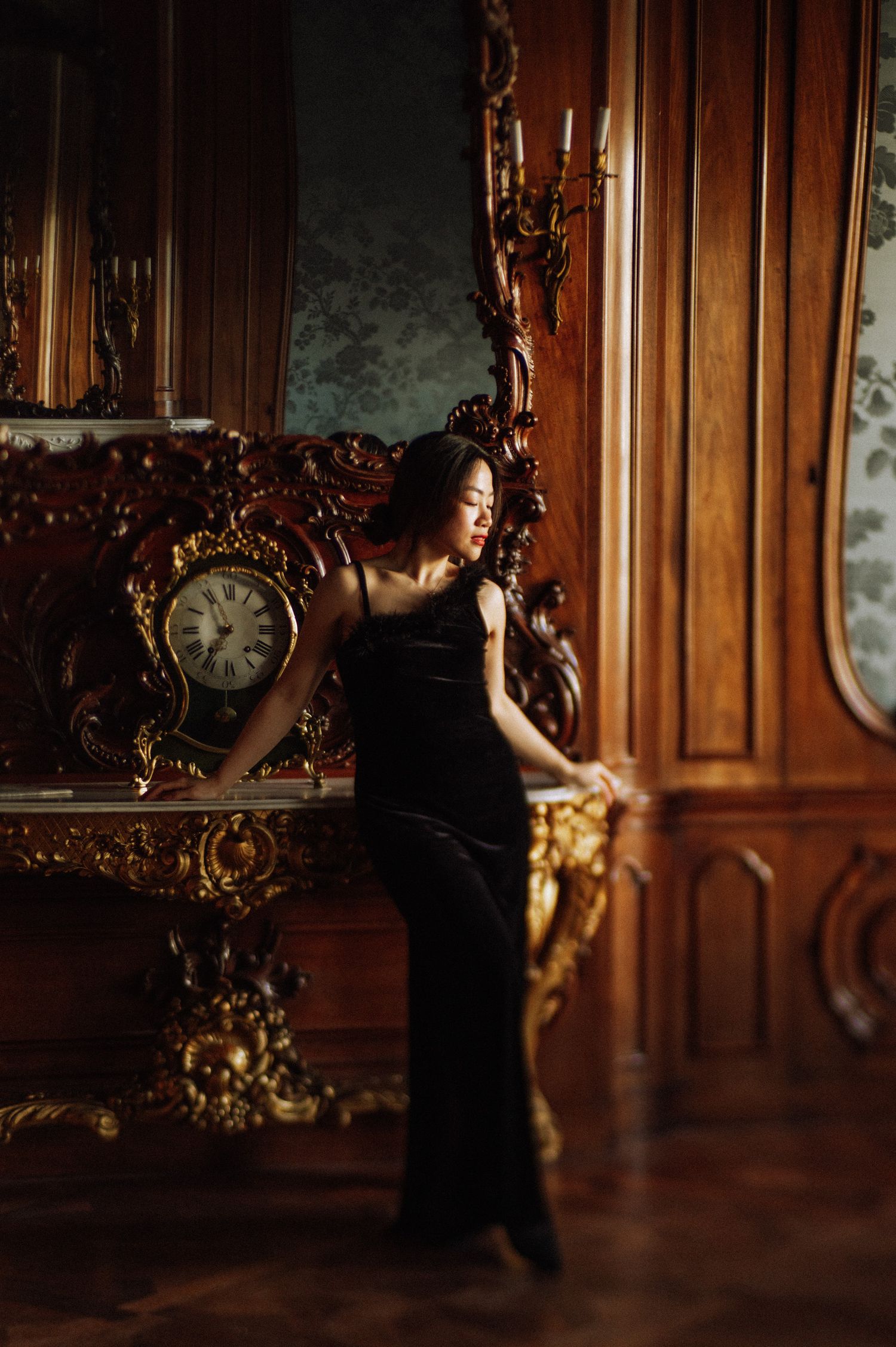 A woman in an elegant black dress poses in a luxurious vintage room with ornate wooden details.
