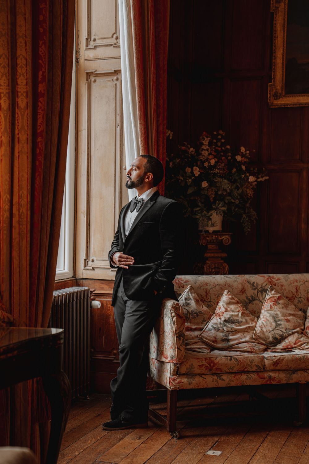 Groom in black tuxedo gazes out tall window in ornate vintage-furnished room at Arley Hall
