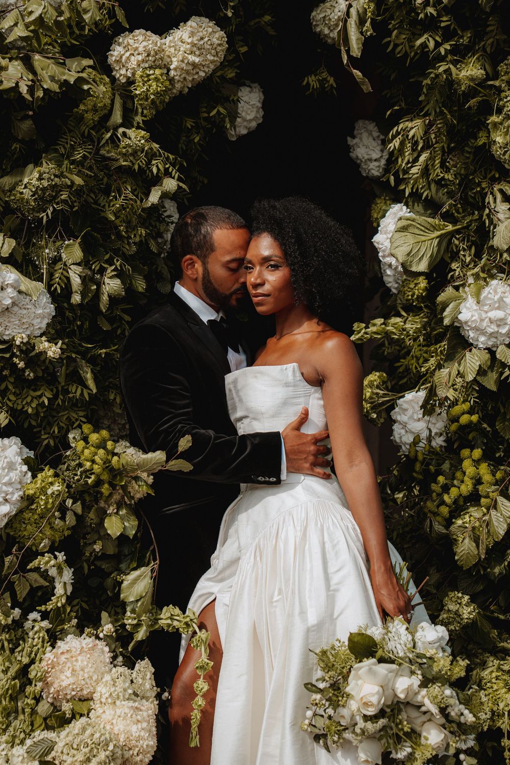 Bride and groom embrace romantically in front of lush white and green floral backdrop at Arley Hall