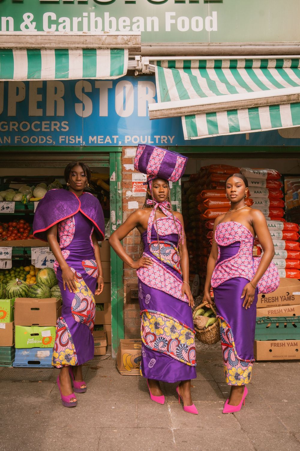 Three women in vibrant purple and pink African print outfits posing outside a Caribbean grocery store.