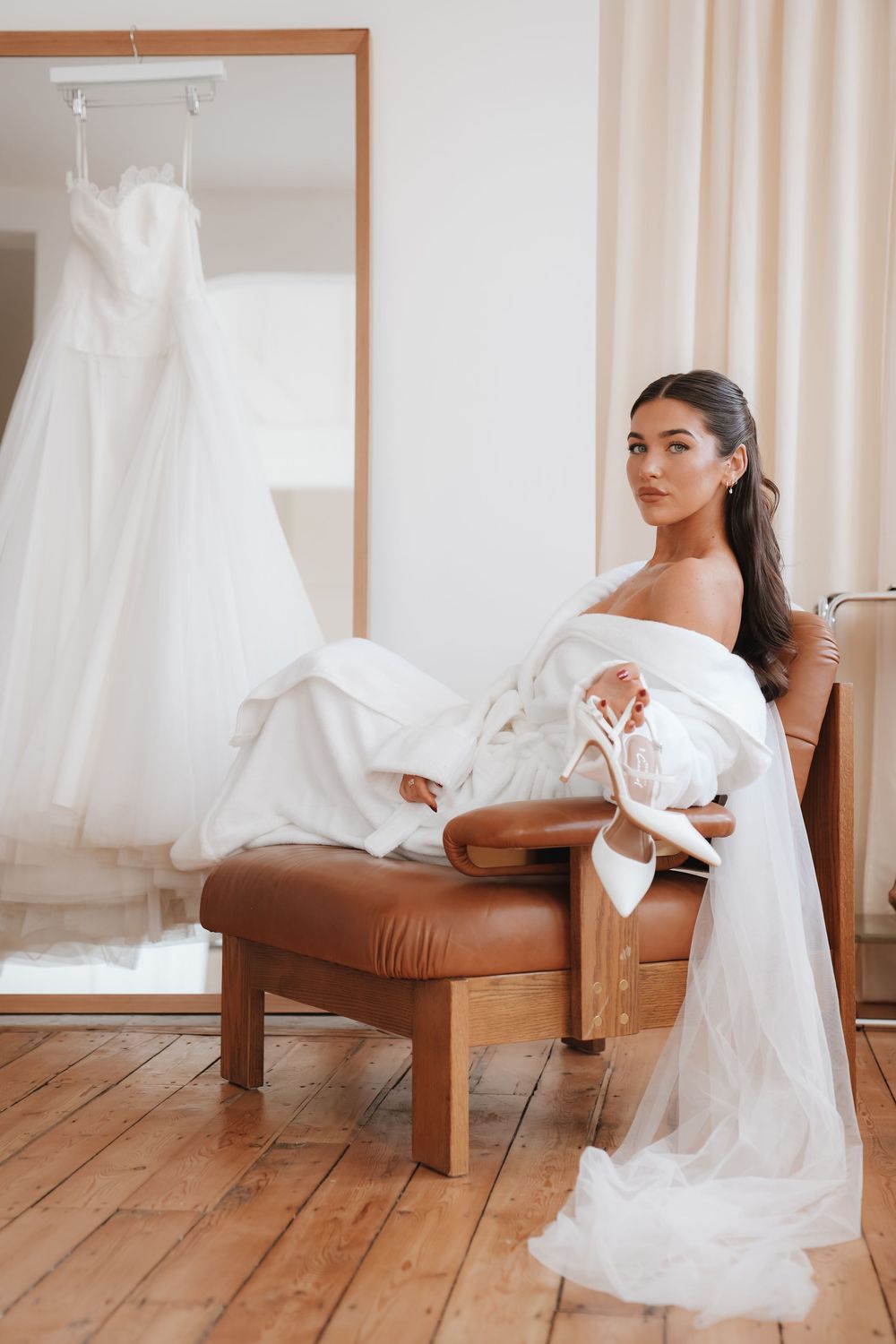 Bride in off-shoulder gown holding heels seated in leather chair with wedding dress reflected.