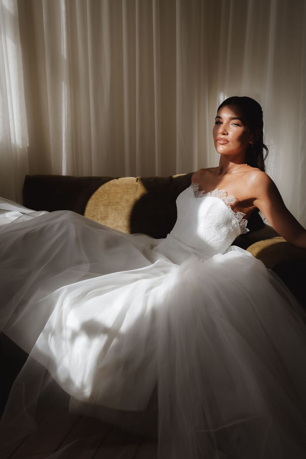 Bride in strapless white tulle ball gown sitting on sofa in soft natural window light.