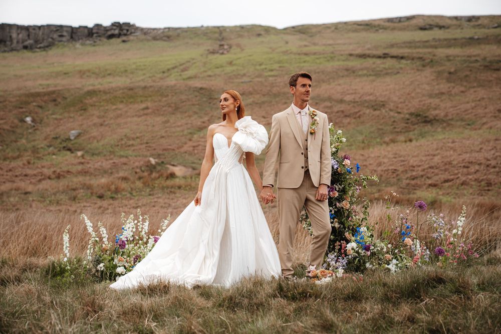 Bride and groom walking hand in hand through a moorland field beside a vibrant wildflower arrangement.