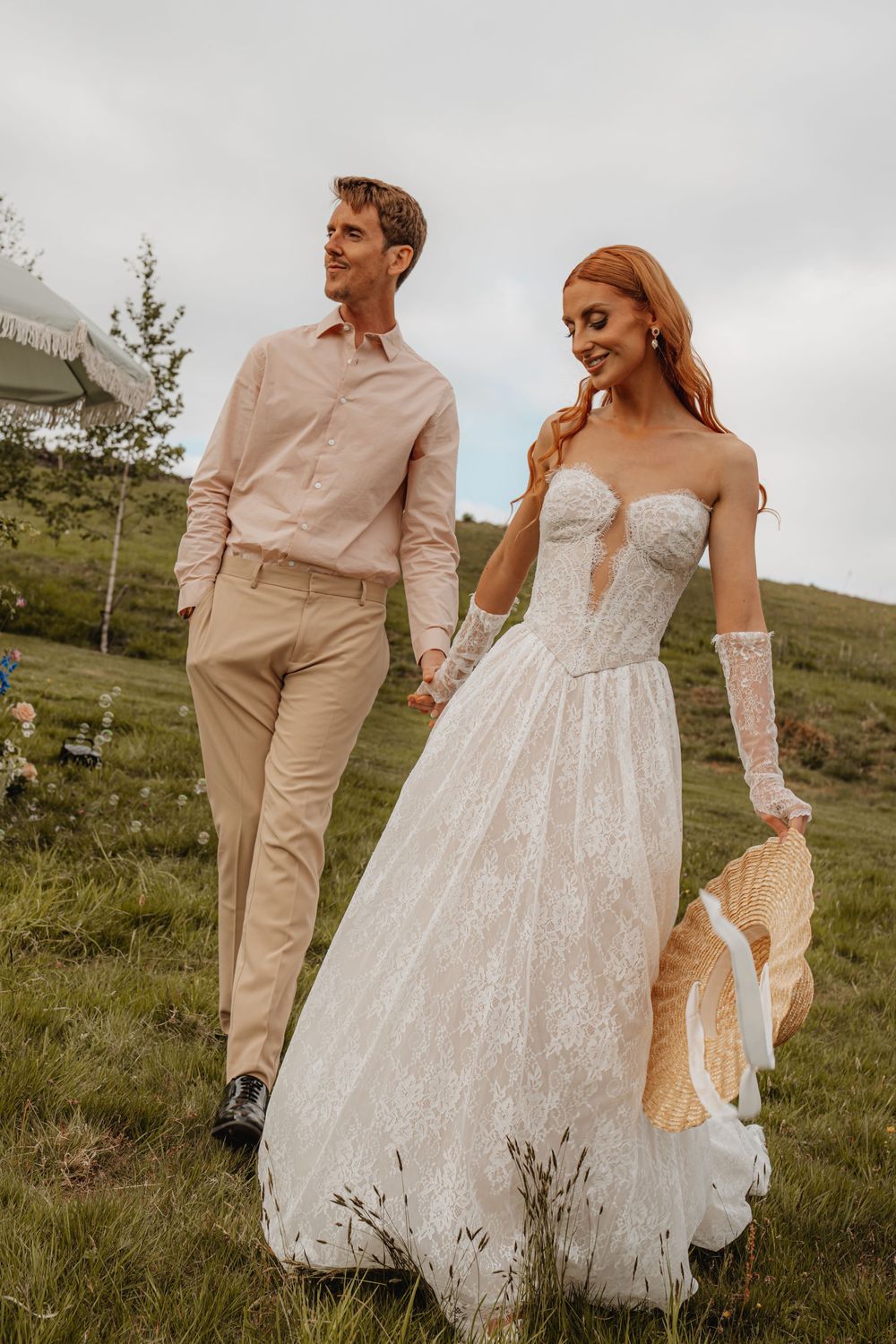 Red-haired bride in a strapless lace corset gown walking with groom through a lush green meadow.