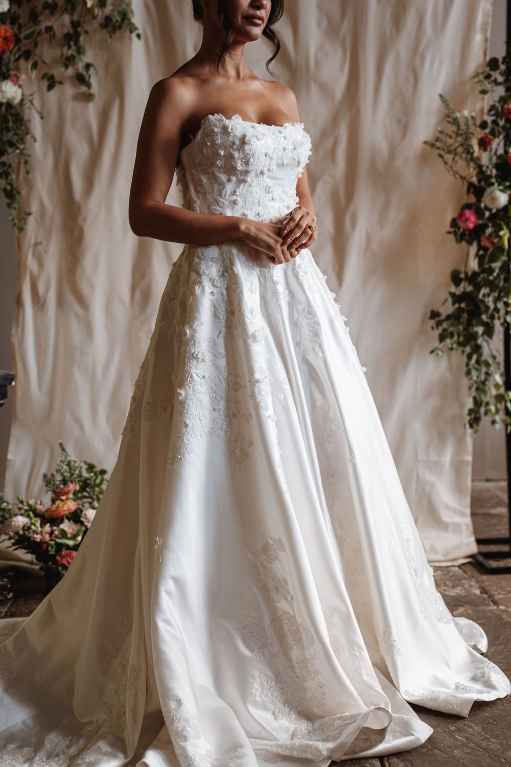 Bride wearing a strapless white ball gown with 3D floral appliques standing before a floral backdrop.