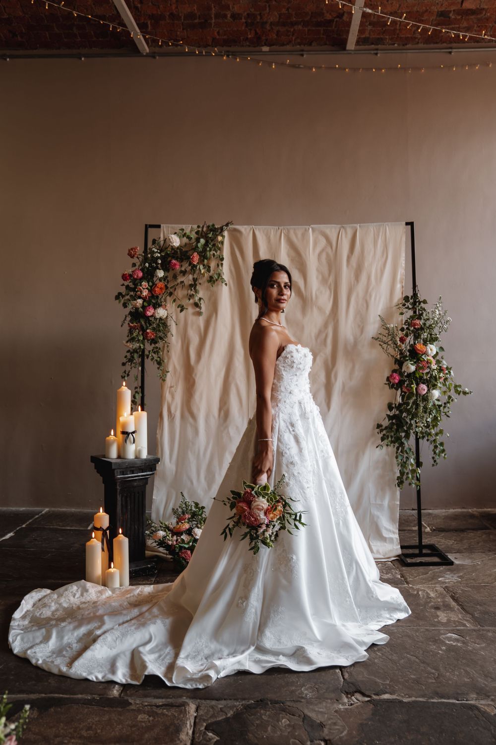 Bride in a strapless lace ball gown standing before a floral backdrop with candles in a rustic venue.
