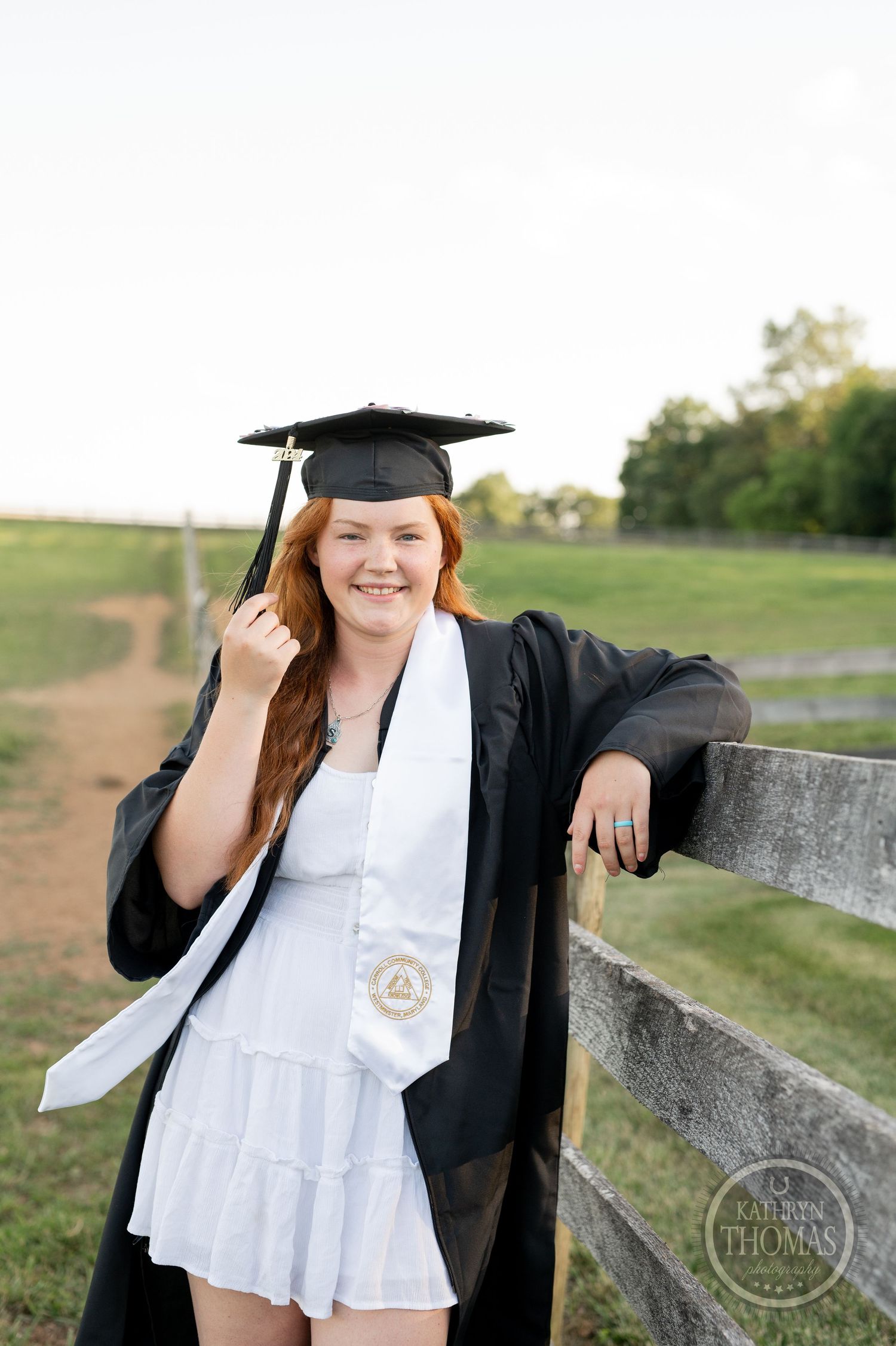 Smiling graduate leans on wooden fence wearing white dress and traditional black graduation regalia.