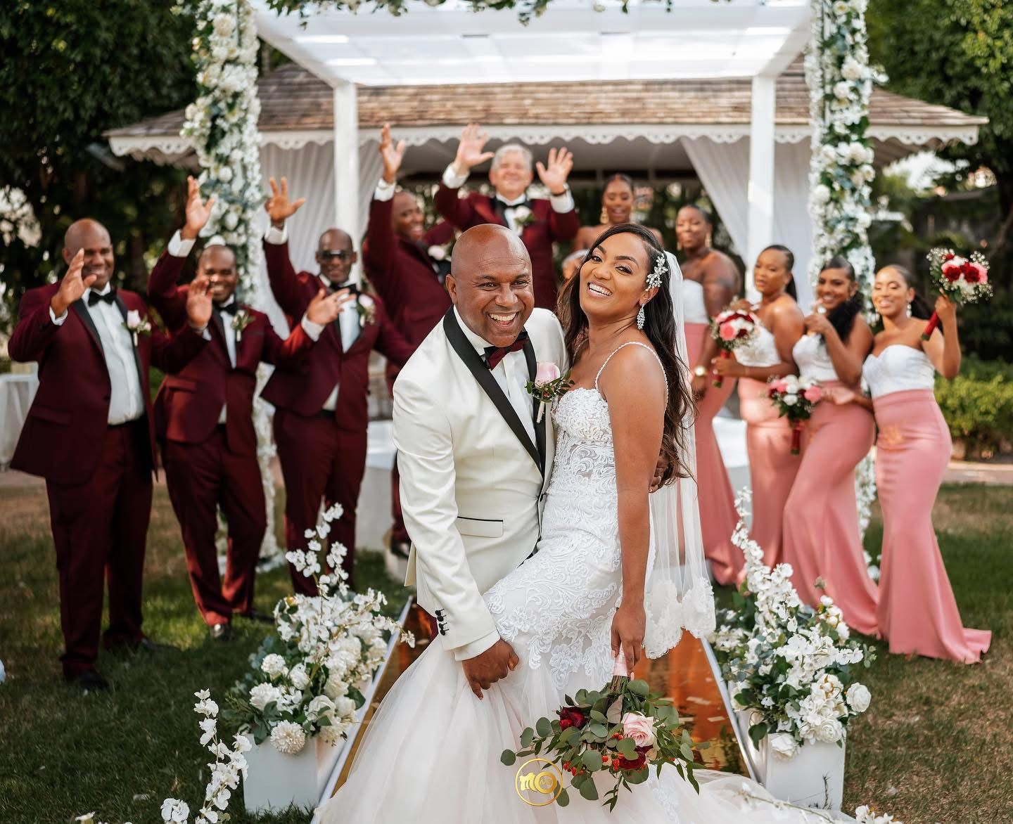Wedding party poses under white floral arch with bridesmaids in pink dresses.