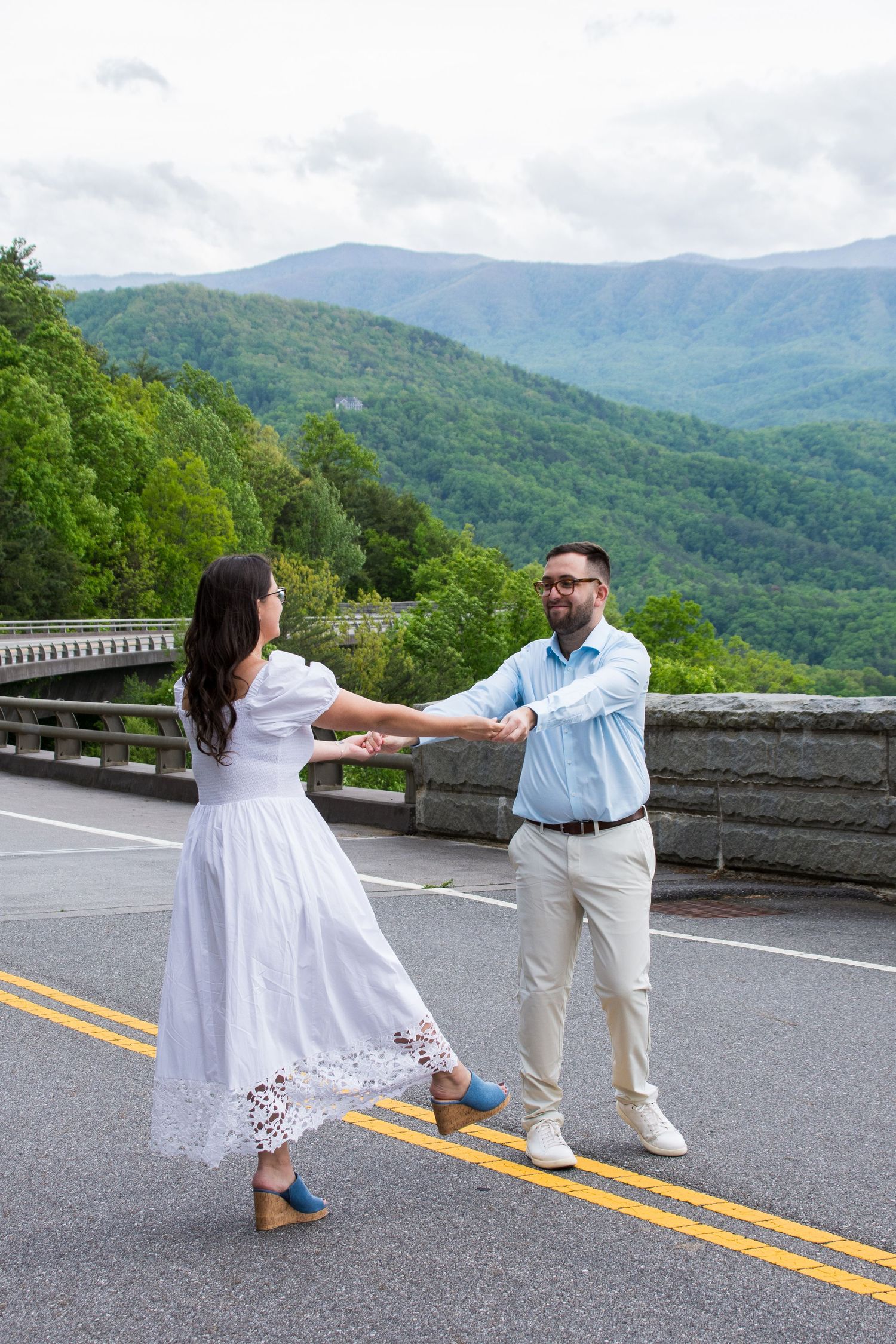 Couple holding hands while hiking along a scenic mountain trail with rolling green hills and forest views in the distance.