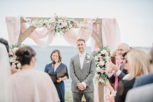 Outdoor wedding ceremony under a blush pink fabric arch decorated with white and pink floral arrangements.