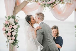 Romantic beach wedding ceremony under a floral arch with pink and white flowers and flowing fabric.