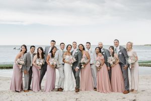 Wedding party in pink dresses and gray suits poses together on a beach under cloudy skies.