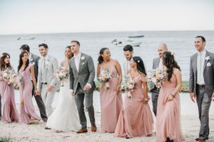 Wedding party in pink dresses and gray suits lined up along the oceanfront for a group photo.