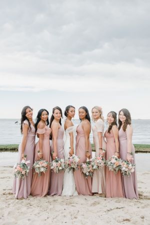 Bridal party in pink and neutral dresses stands in a line on beach holding pastel flower bouquets.