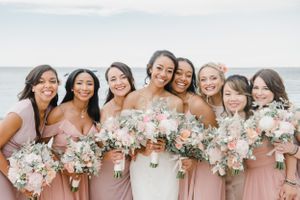 Wedding party in blush pink bridesmaid dresses pose together with elegant floral bouquets by the ocean.