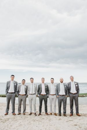 A wedding party of groomsmen in matching gray suits poses on a cloudy beach.