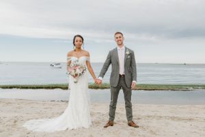 A wedding couple poses hand-in-hand on a beach with a cloudy coastal backdrop.
