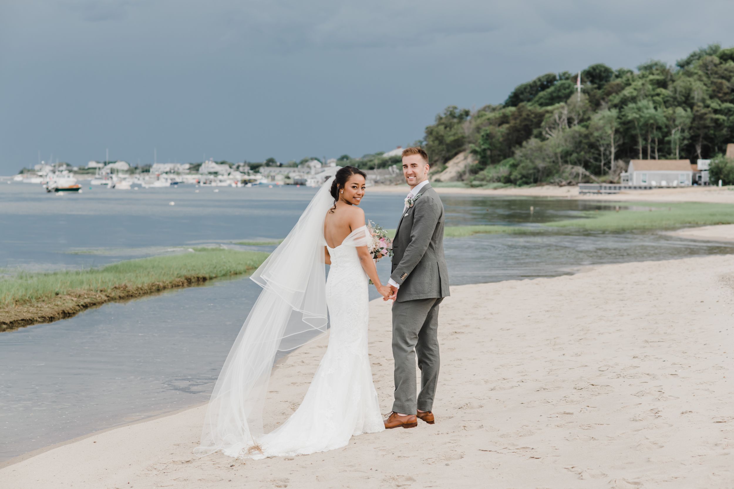 A couple in wedding attire stands together on a sandy beach with boats visible in the distance.