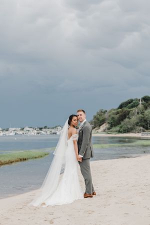 A newlywed couple poses on a scenic beach with boats and greenery in the background.