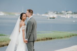 A wedding couple shares a tender moment on a beach with boats in the harbor behind them.