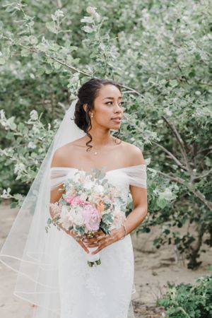 Bride in off-shoulder gown holding pink and peach bouquet against flowering bush.
