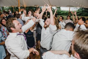 Wedding reception guests dancing and celebrating under a tent with their hands in the air.