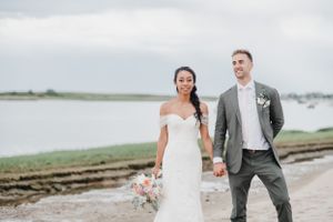 A joyful couple in wedding attire pose together on a coastal path with water and boats in the background.