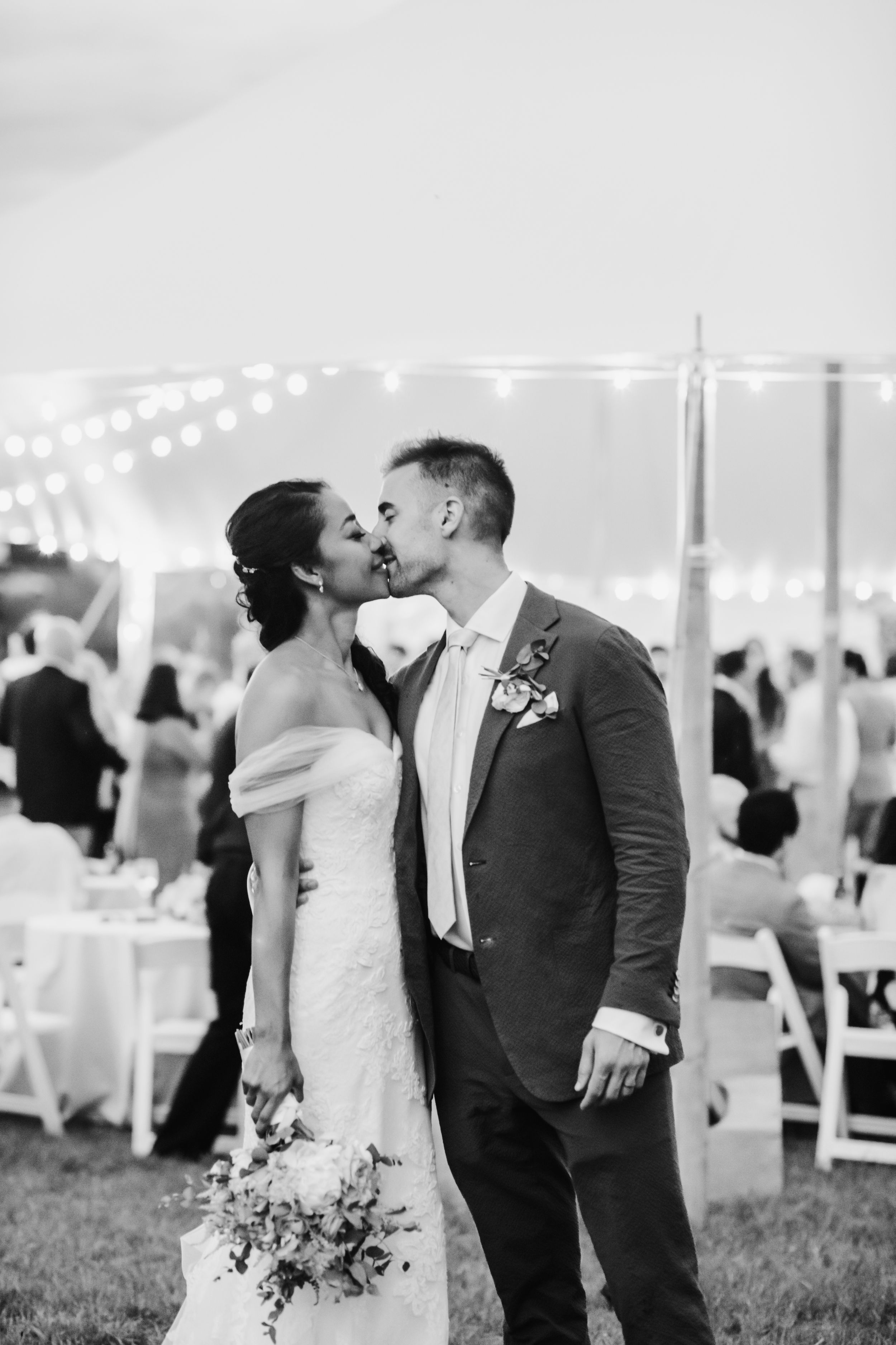 A romantic black and white wedding moment under a tent with string lights and white tables in the background.
