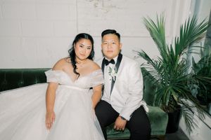 Formal couple poses together on green velvet couch with tropical palm decor at elegant wedding reception.
