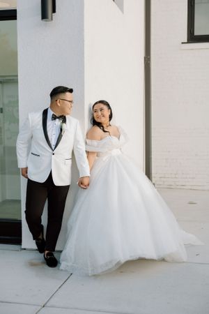 A couple in formal wedding attire walks together outside a white building with glass windows.
