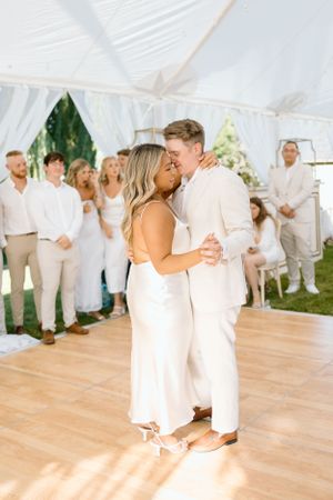 A wedding couple shares their first dance under a white tent while wedding guests watch from the background.