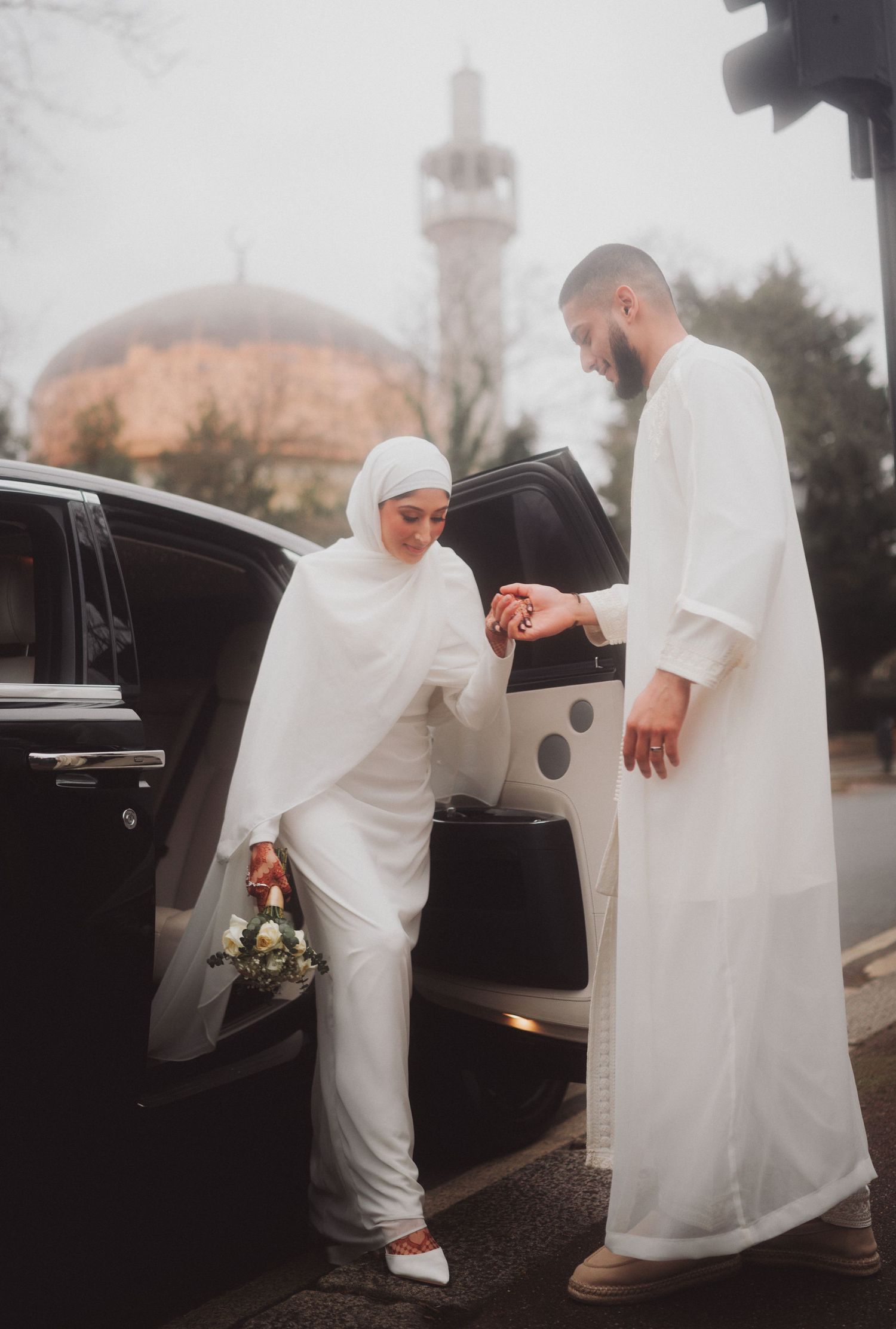 Traditional Muslim couple in white attire by a car with mosque in background.