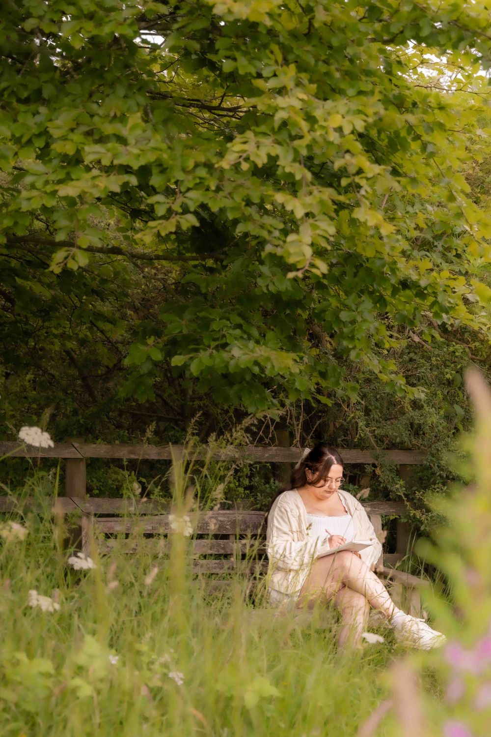 Person in white dress sitting on rustic bench surrounded by tall grass and green foliage. Lincolnshire branding photographer.