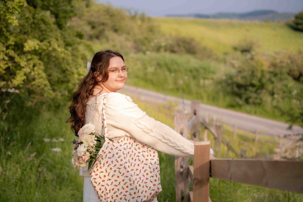 Figure in cream sweater and floral dress leaning against wooden fence with countryside view. Lincolnshire branding photographer.