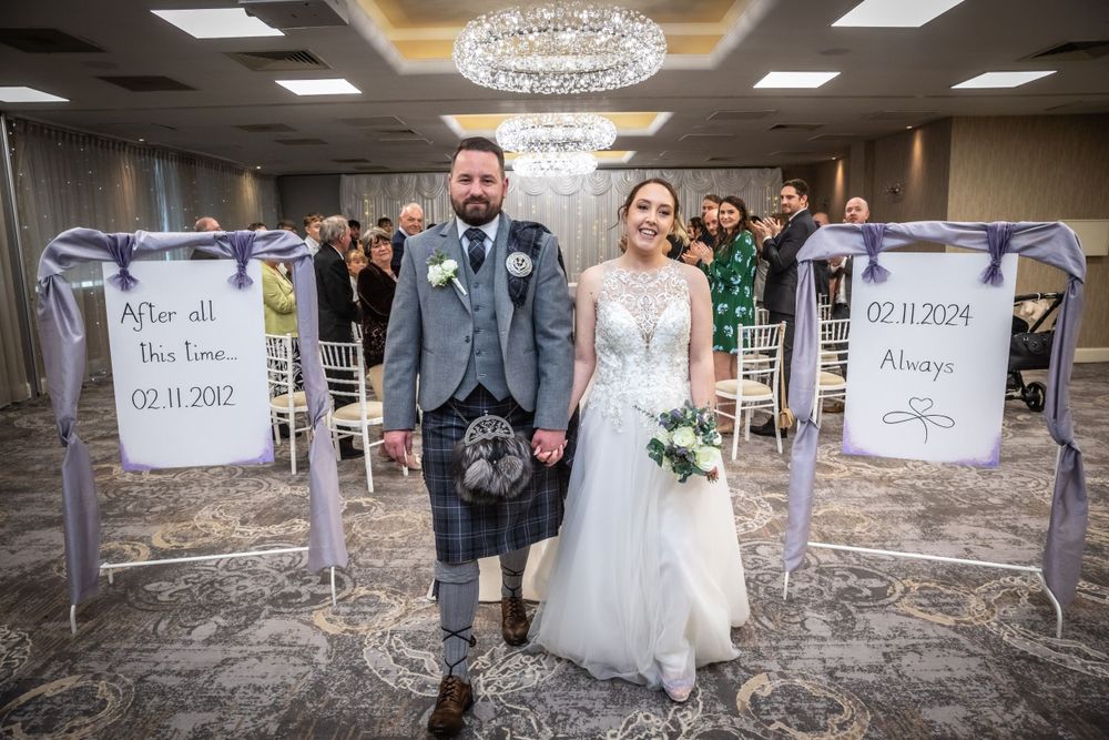 Bride and groom walking down the aisle in an elegant ballroom decorated with purple wedding signs.