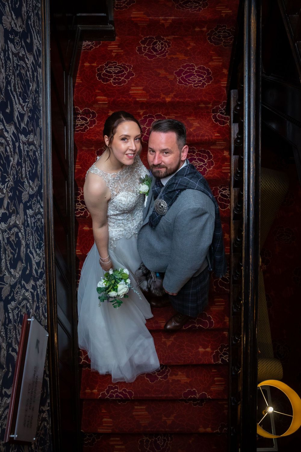 Bride and groom posing together on a grand staircase with ornate red carpet.