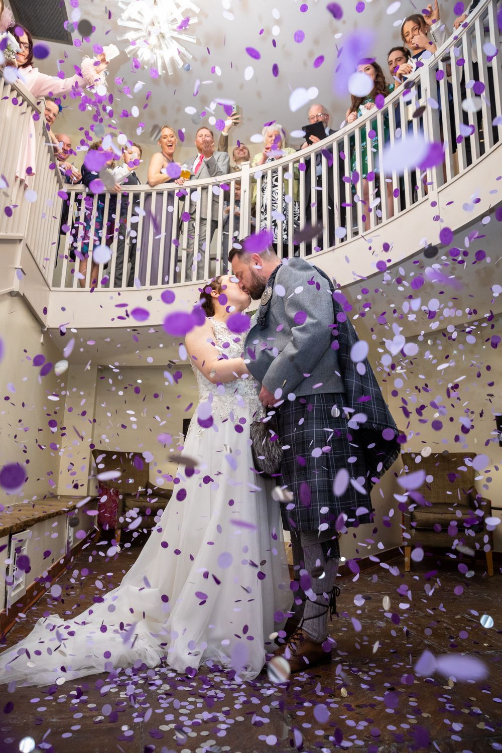Bride and groom sharing a kiss beneath a shower of purple confetti on a grand staircase.