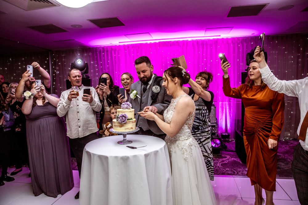 Bride and groom cutting their wedding cake surrounded by cheering guests under pink lighting.