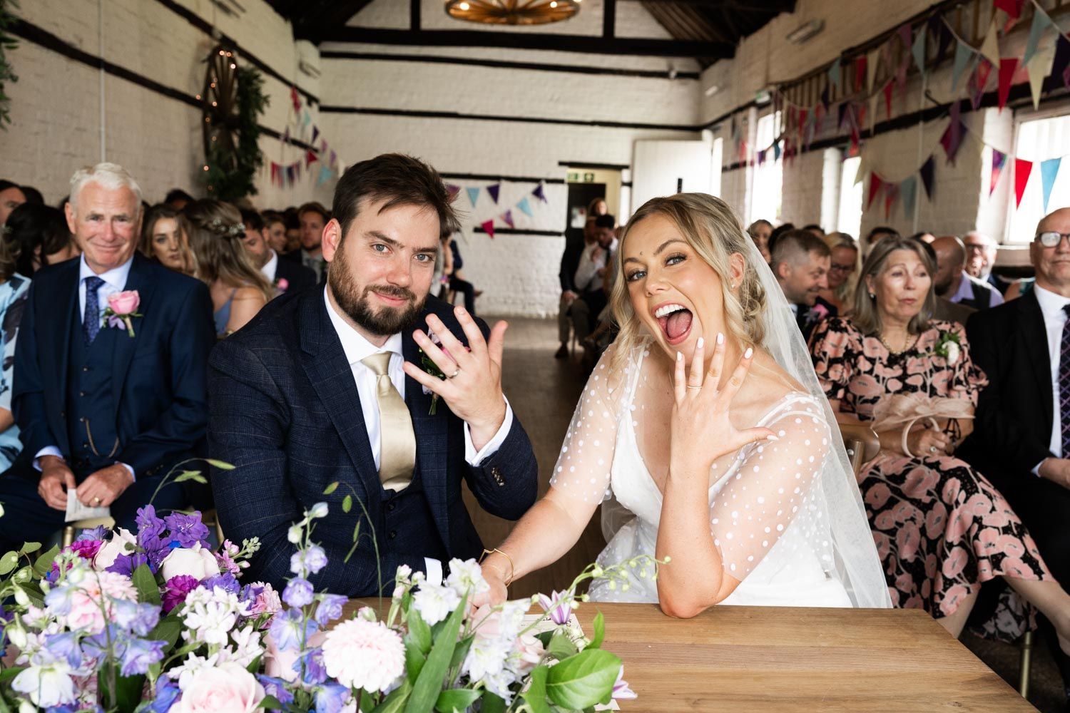 Two people share a lighthearted, candid moment during their wedding ceremony as they sign documents at a decorated table.
