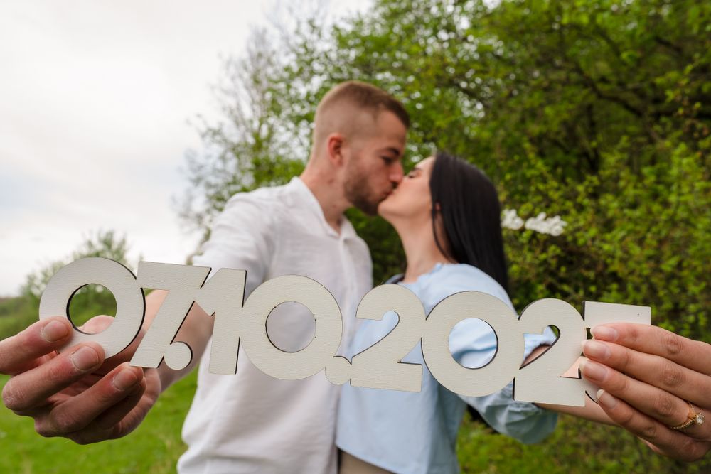 Couple holding save the date numbers in a field with green trees in background.