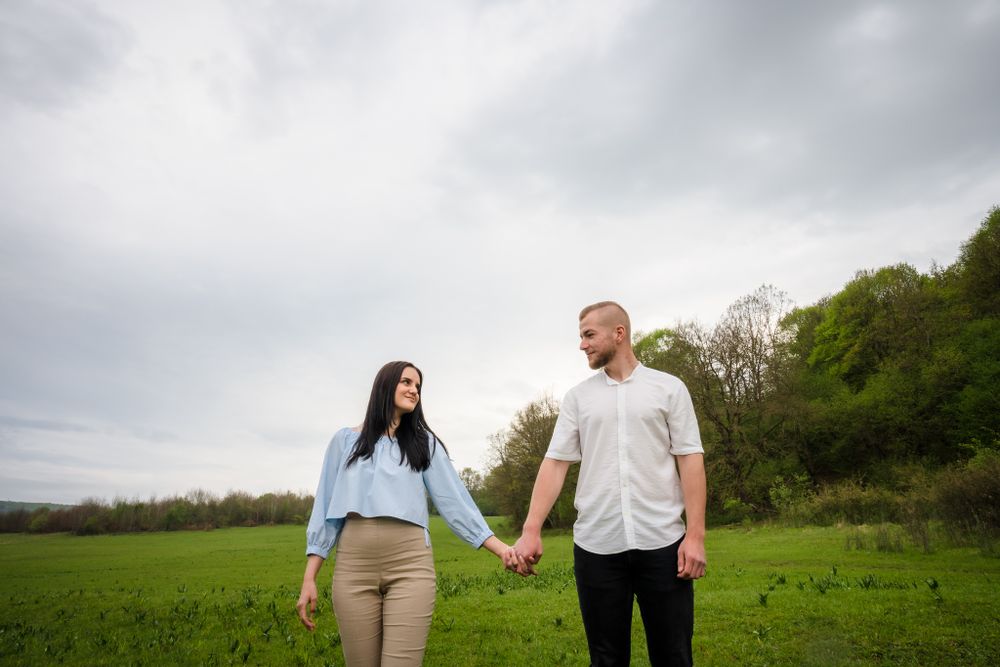 Couple walking hand in hand across a green field under cloudy skies.