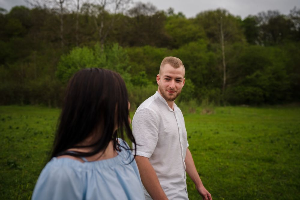Couple posing together in a scenic field with trees and cloudy sky backdrop.