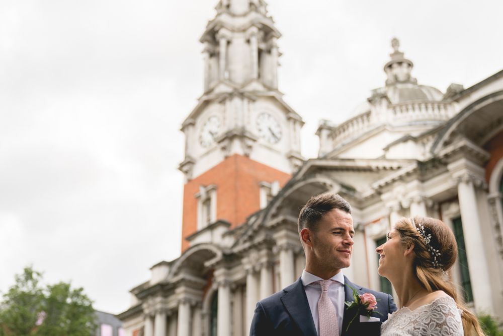 Couple in wedding attire pose outside historic building with ornate clock tower.