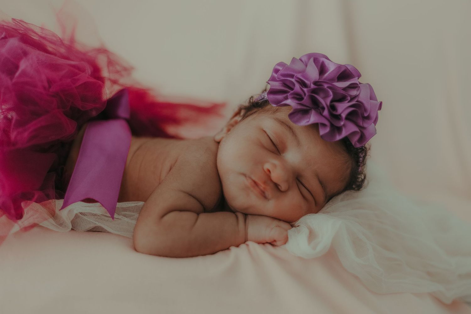 A sleeping baby wearing a purple flower headband and pink tulle wrap.