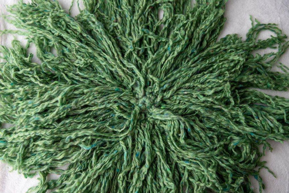 Round green curly yarn fiber bundle spread flat on white surface.